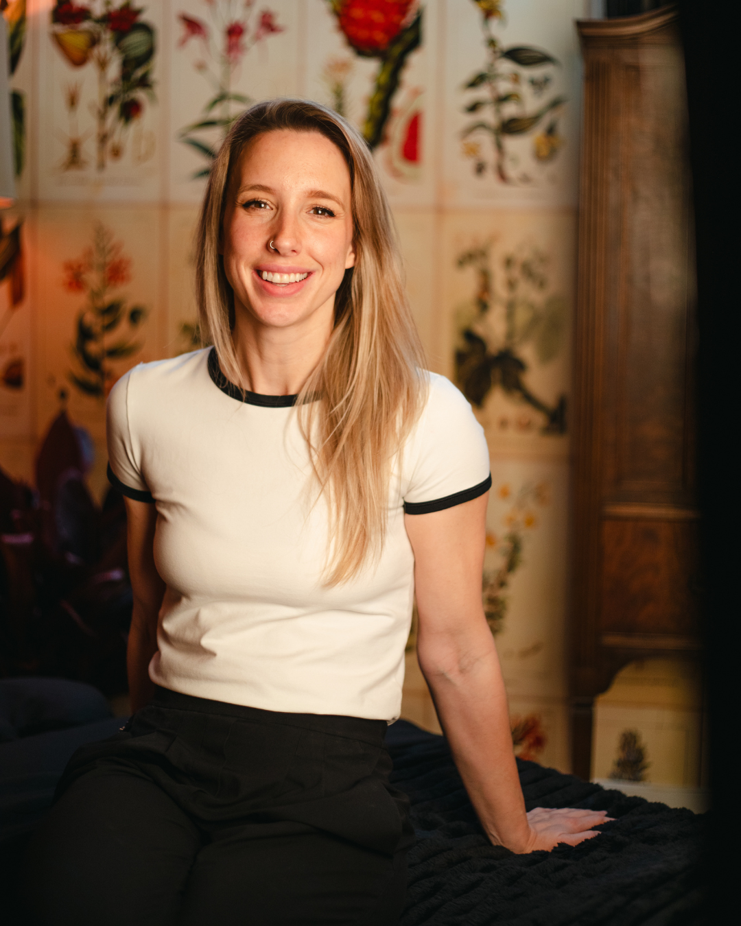 A smiling woman with blonde hair wearing a black top standing outdoors with mountains and city in the background during sunset.