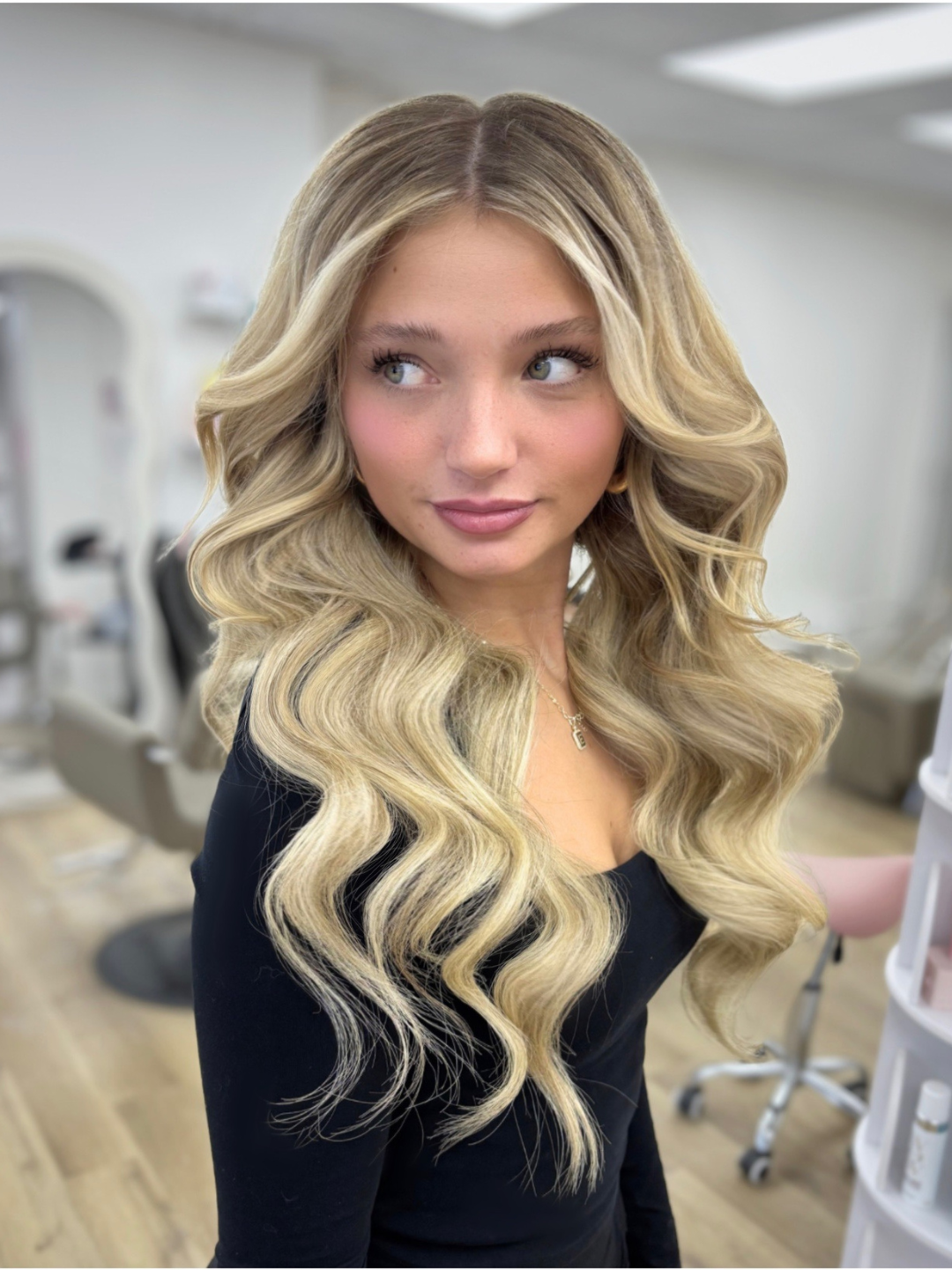A young woman with long, wavy blonde hair, wearing a black top and a gold necklace, standing in a salon or spa setting.
