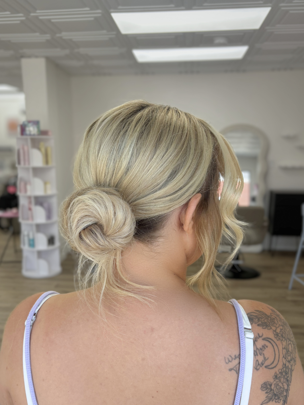 A woman with blonde hair styled in an elegant low bun with a loose curl framing her face, in a salon or hair studio.