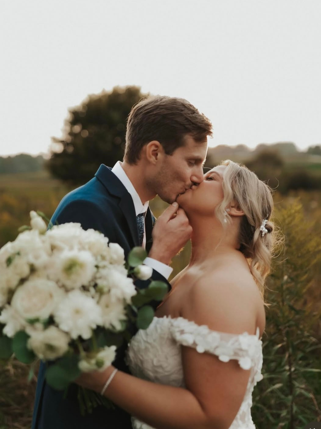 A newlywed couple sharing a kiss outdoors, with a bouquet of white flowers, during sunset or late afternoon.
