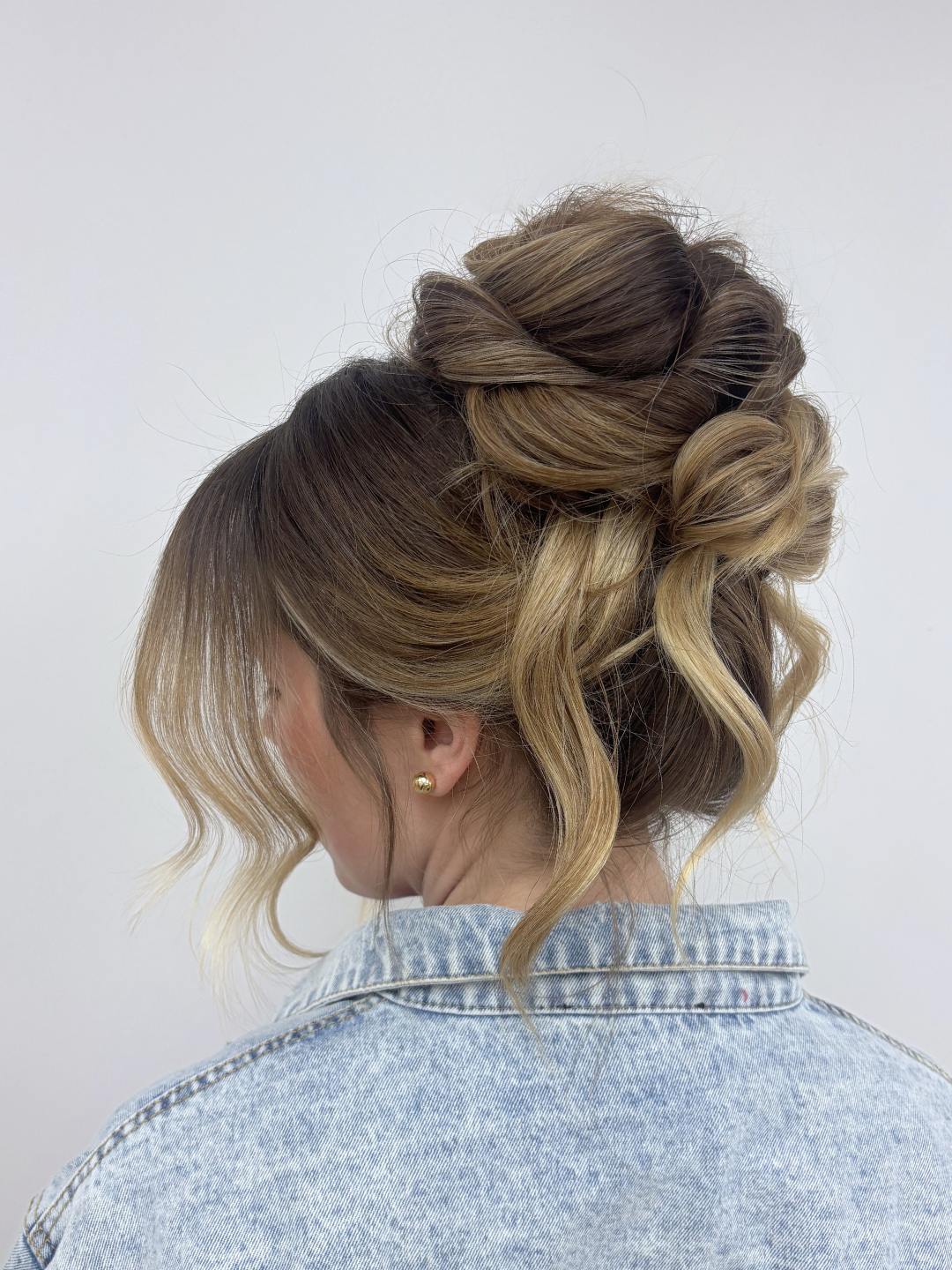 A woman with styled hair, wearing a denim jacket and pearl earrings, seen from behind against a plain white background.