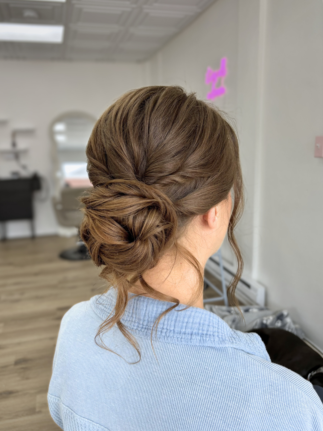 Back view of a woman with styled brown hair in an elegant updo with loose curls, wearing a light blue top, in a salon or studio setting.