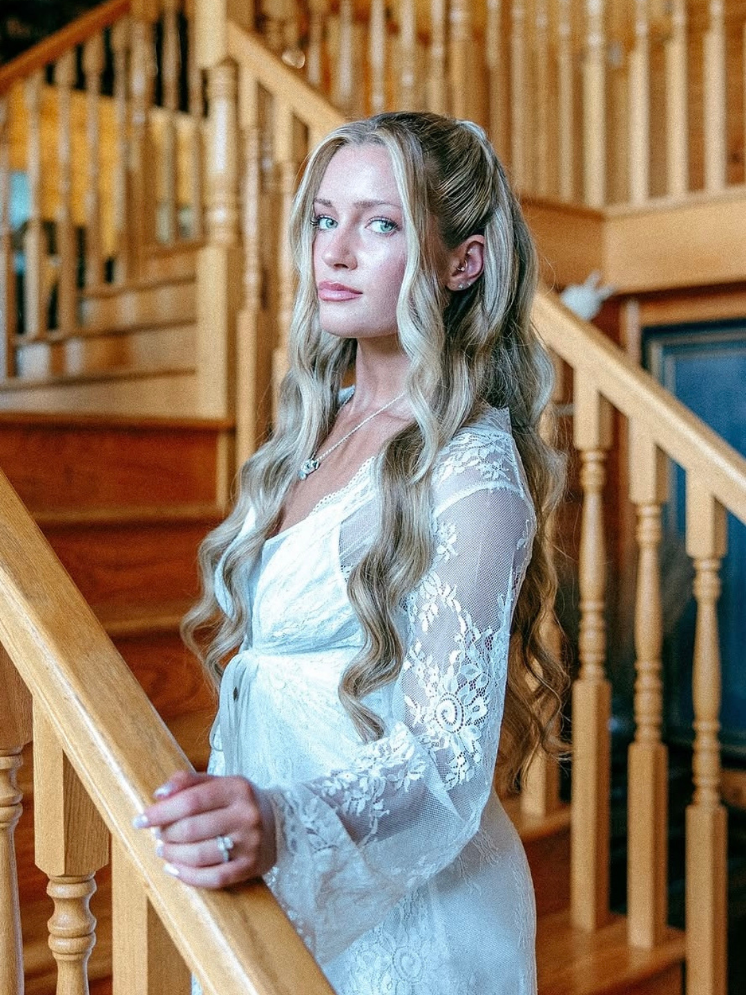 A woman with long, wavy blonde hair standing on a wooden staircase inside a house, wearing a white lace dress and a necklace, looking at the camera.