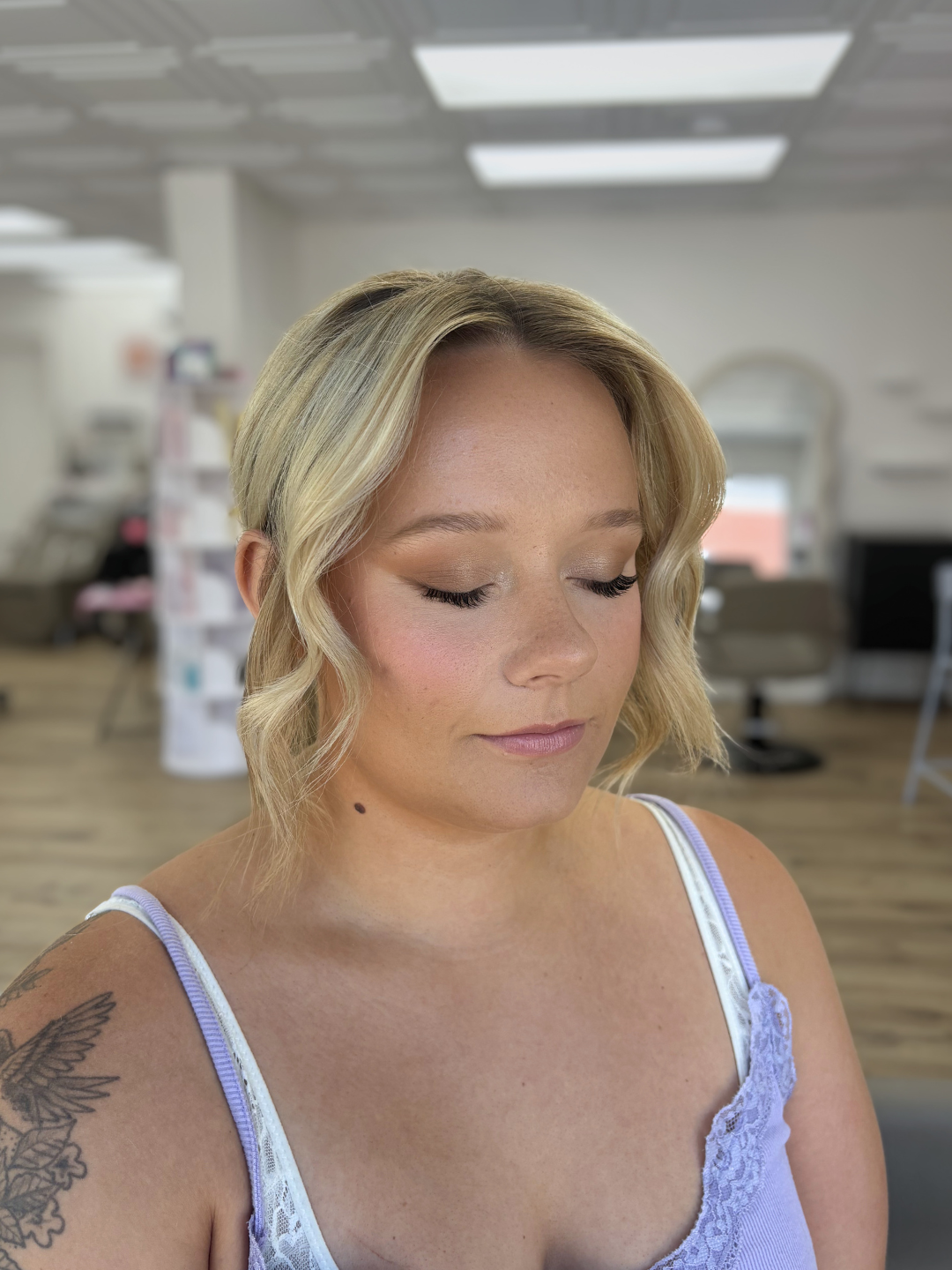 A woman with blonde wavy hair and a tattoo on her left shoulder, wearing a lavender and white lace top, with her eyes closed, in a salon or spa setting.