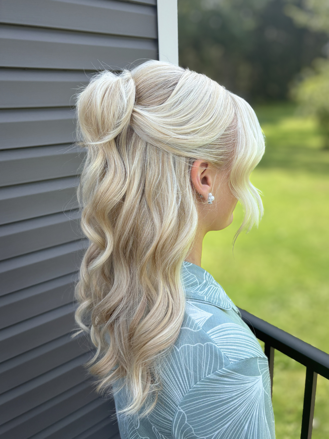 Blonde woman with wavy hair styled in a half-updo, standing outdoors near a gray house wall and a black railing, with greenery in the background.