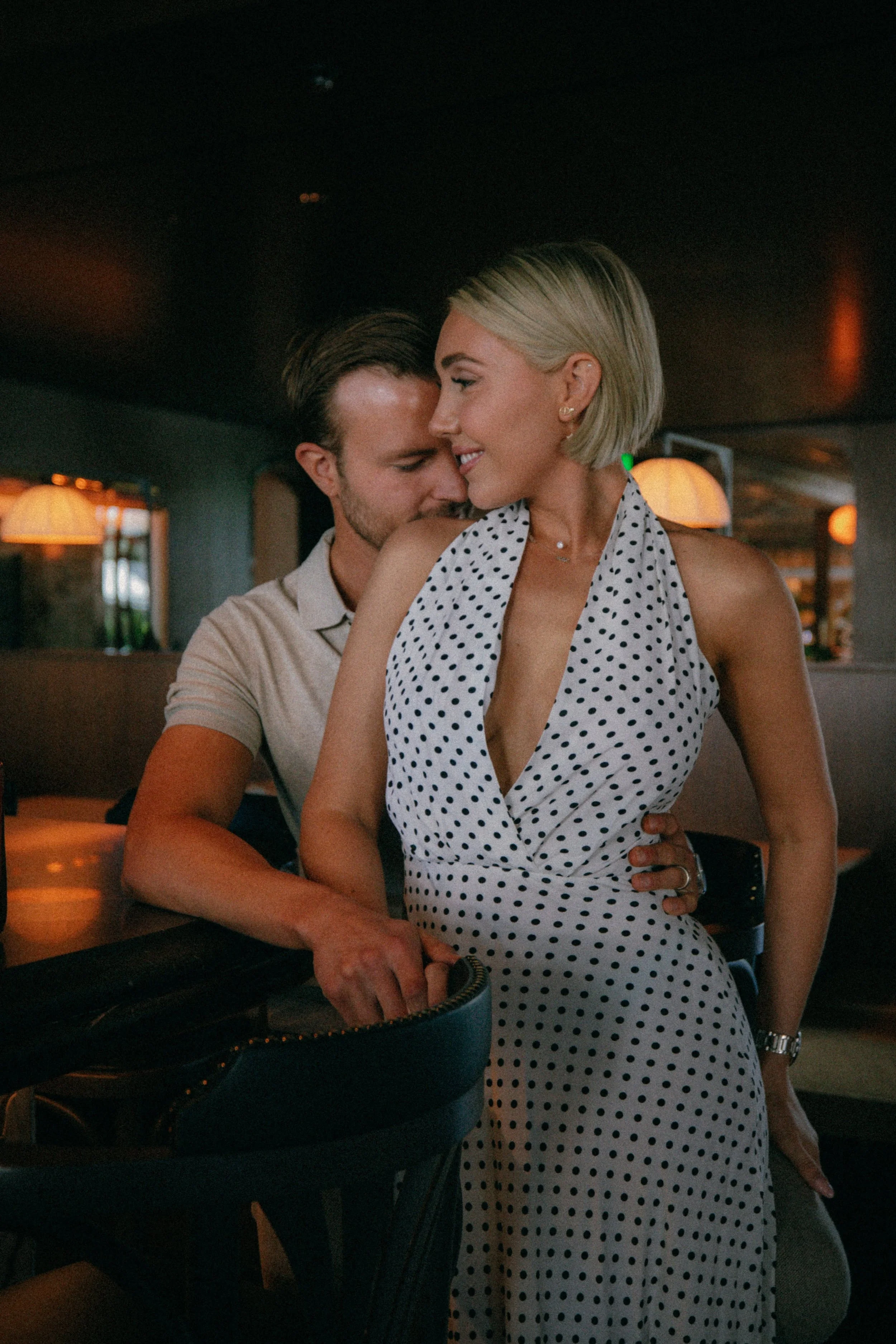 A man and woman sharing an intimate moment in a dimly lit restaurant or bar, with the man close behind and touching the woman's waist, both smiling.