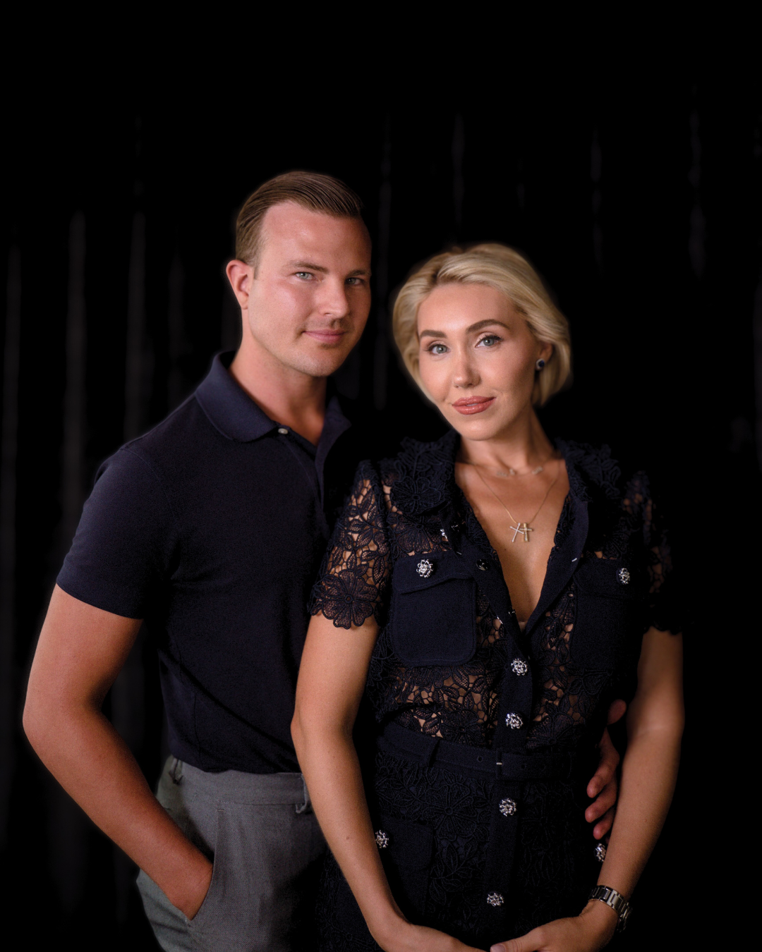 A man and woman posing together against a black background. The man is wearing a dark polo shirt and light gray trousers, and the woman is dressed in a black lace top with embellishments, with blonde hair styled in loose waves.