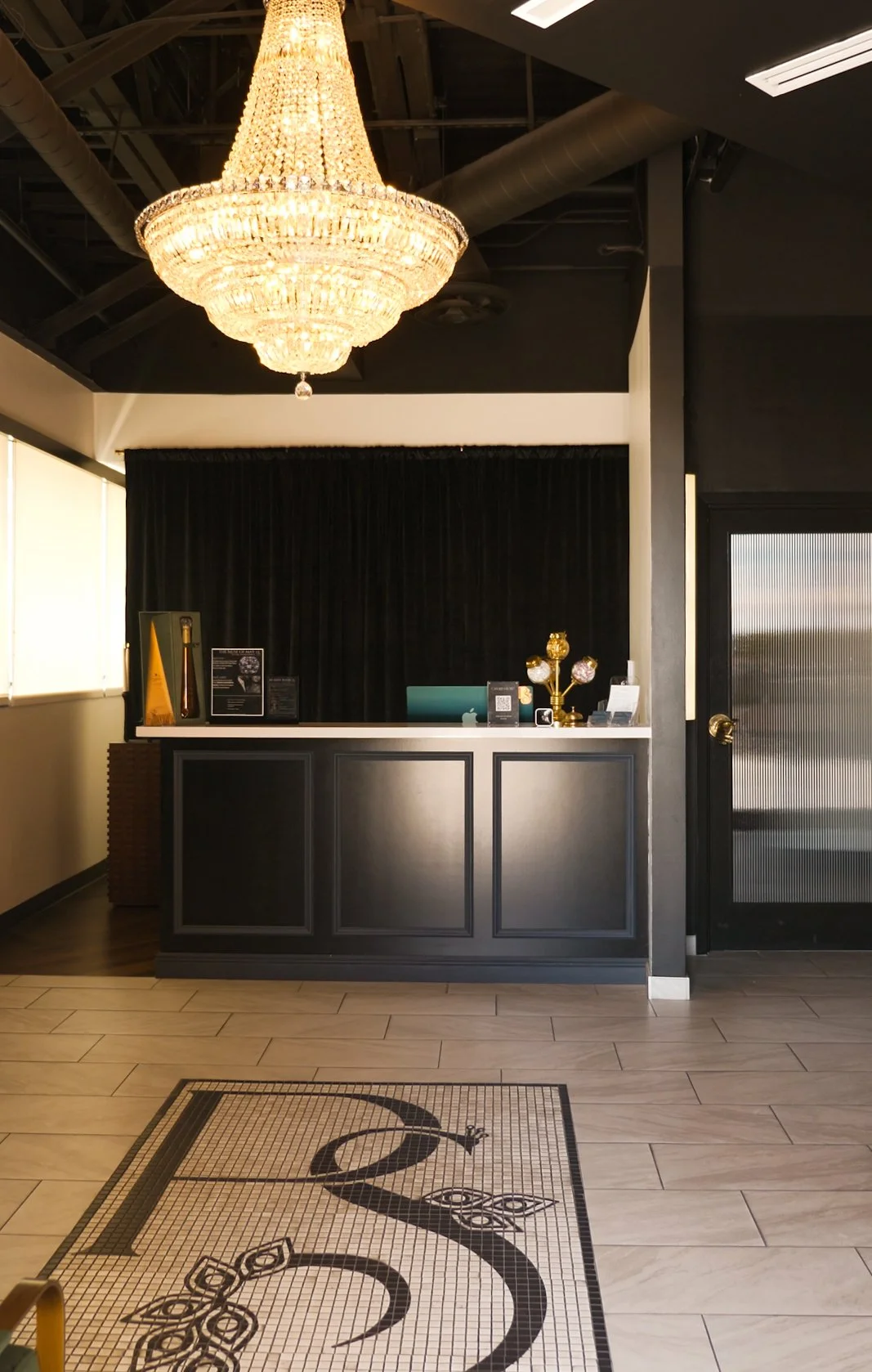 Luxurious hotel lobby reception area with a large crystal chandelier, black curtained reception desk with decorative gold and glass fixtures, and a tiled floor featuring a stylized black logo.