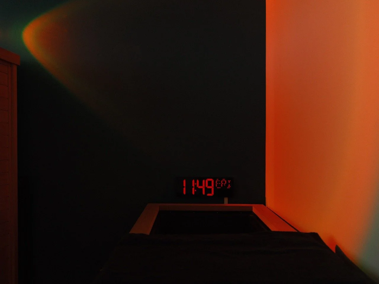 A digital clock on a wall shows the time as 11:49, with a dark room illuminated by a soft orange light on the right side and a faint rainbow-colored light on the left side.