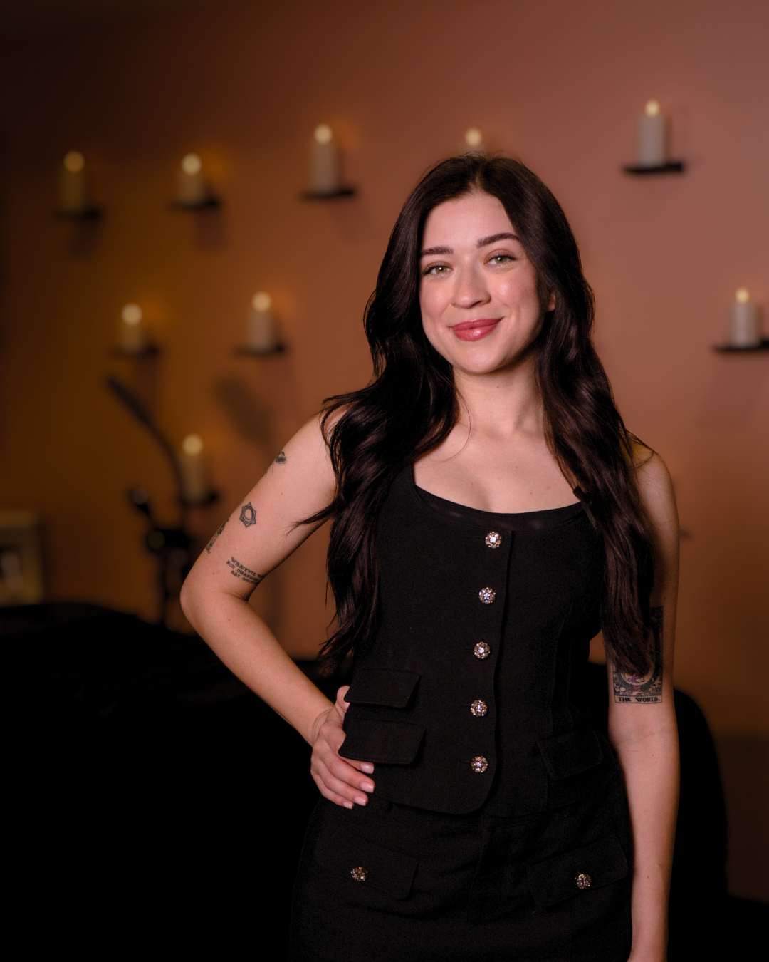 A young woman with long dark hair, wearing a black sleeveless dress with decorative buttons, standing indoors with a warm-colored wall and candle wall sconces in the background.