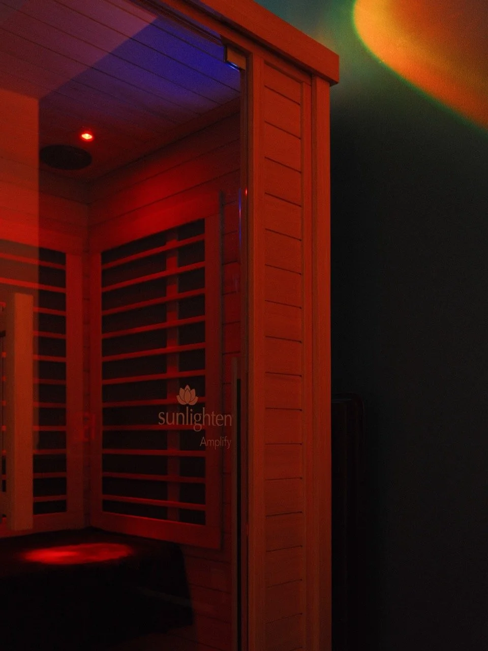 An interior of a wooden sauna room illuminated with red and green lights, featuring wooden benches and a door with the 'sunlighten Amplify' logo.