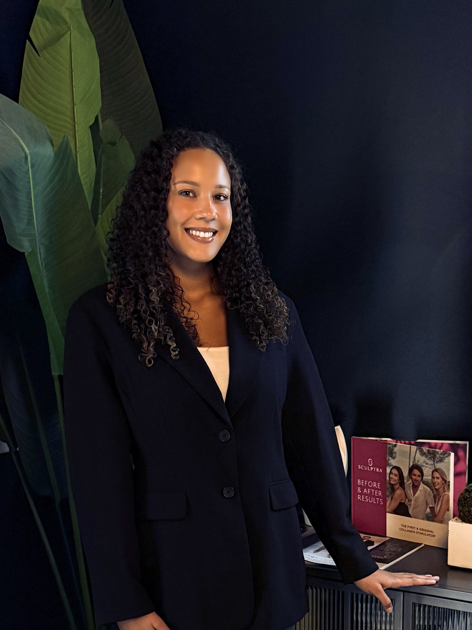 A woman with curly hair in a black blazer and white shirt standing next to a table with brochures and a model of a human head. The background features large green leaves and a dark blue wall.