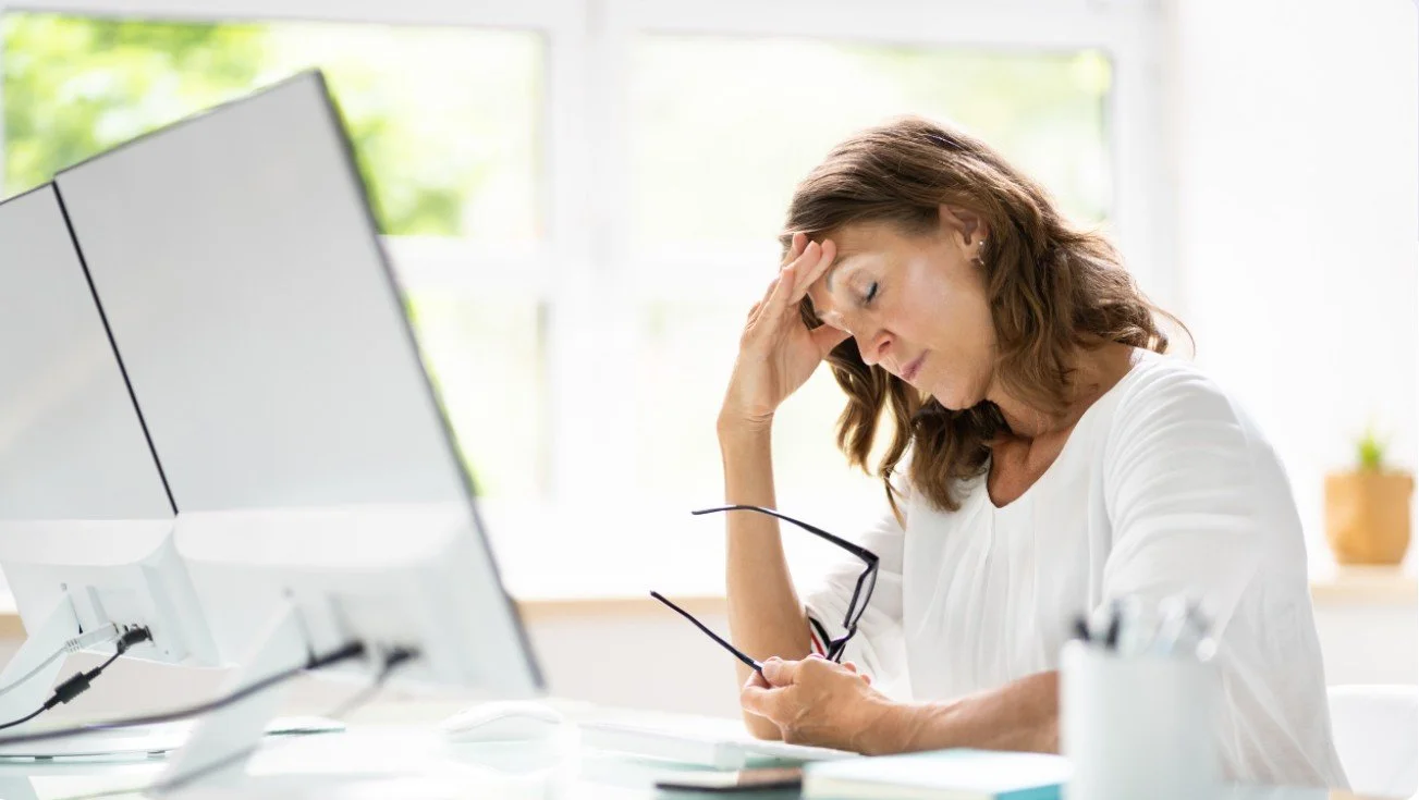 A woman sitting at a desk holding her forehead with her left hand and glasses in her right hand, appearing stressed or tired, with a computer monitor in front of her.