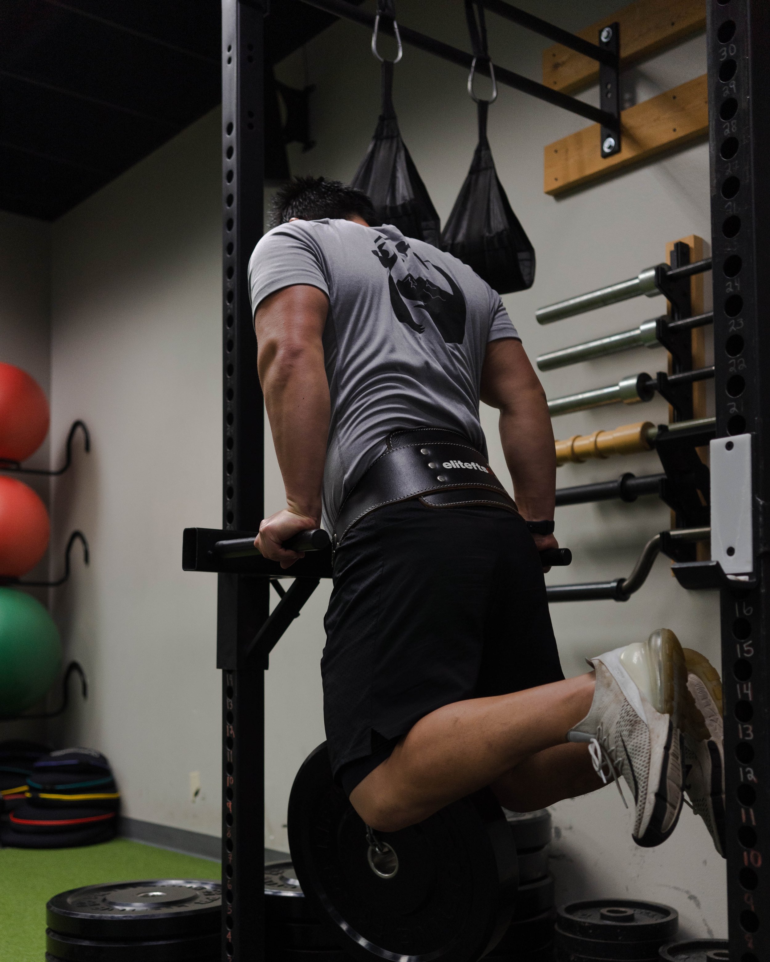 A man doing a dip exercise on parallel bars in a gym, facing away from the camera, with weight plates and gym equipment visible around him.