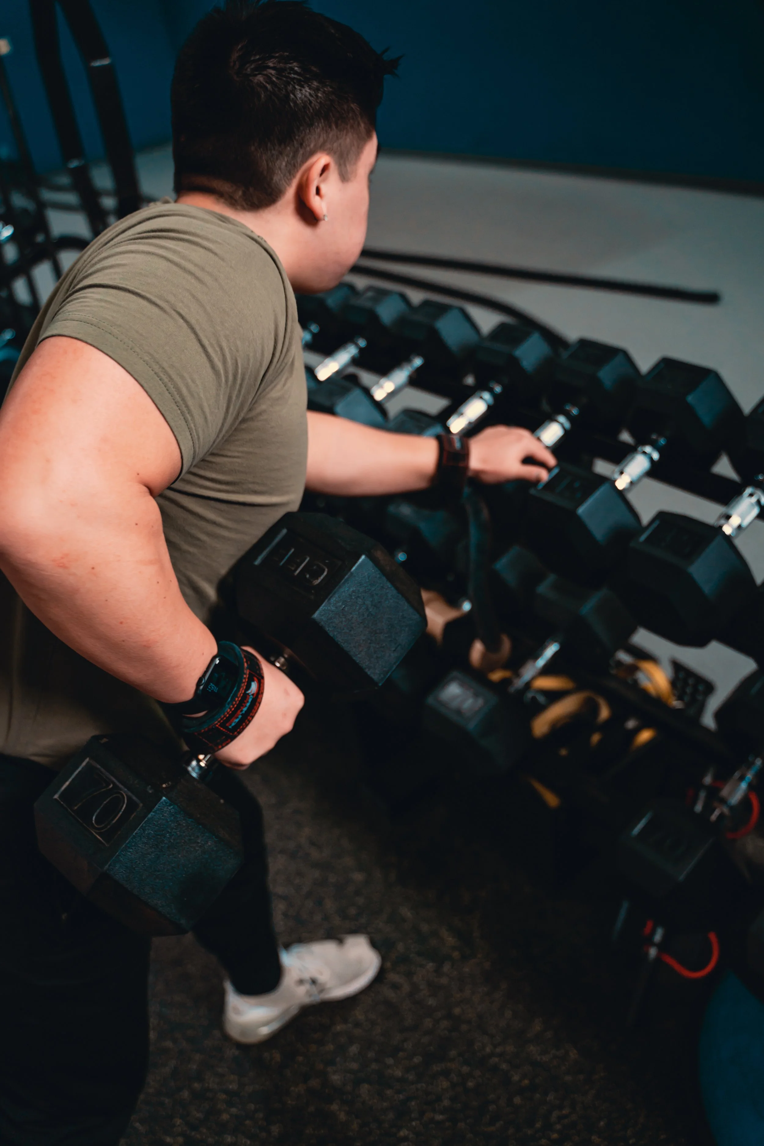 Person lifting a dumbbell in a gym, with a row of dumbbells on a rack in front of them.