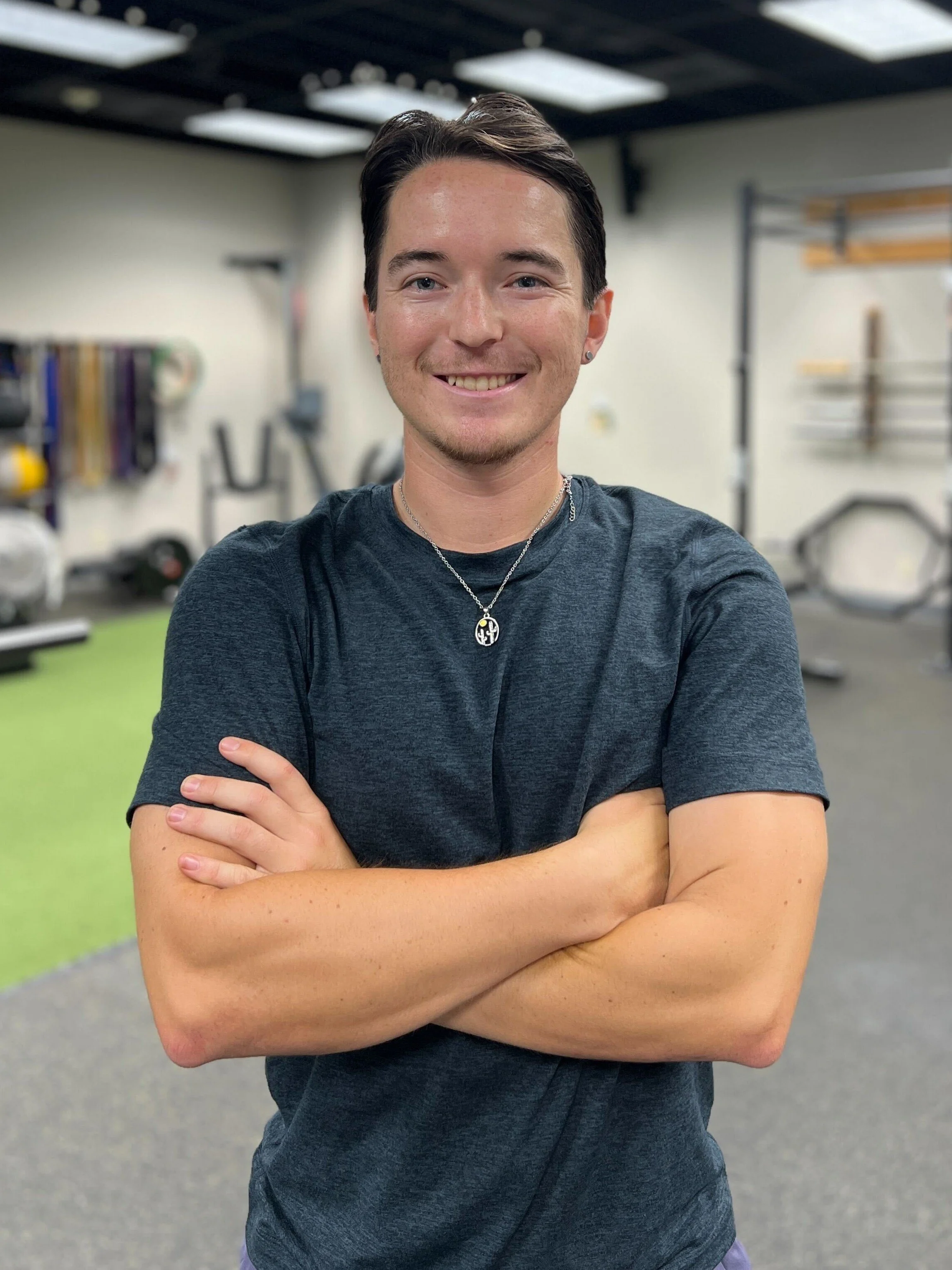 A young man with dark hair and light skin smiling and crossing his arms in a gym or fitness center.