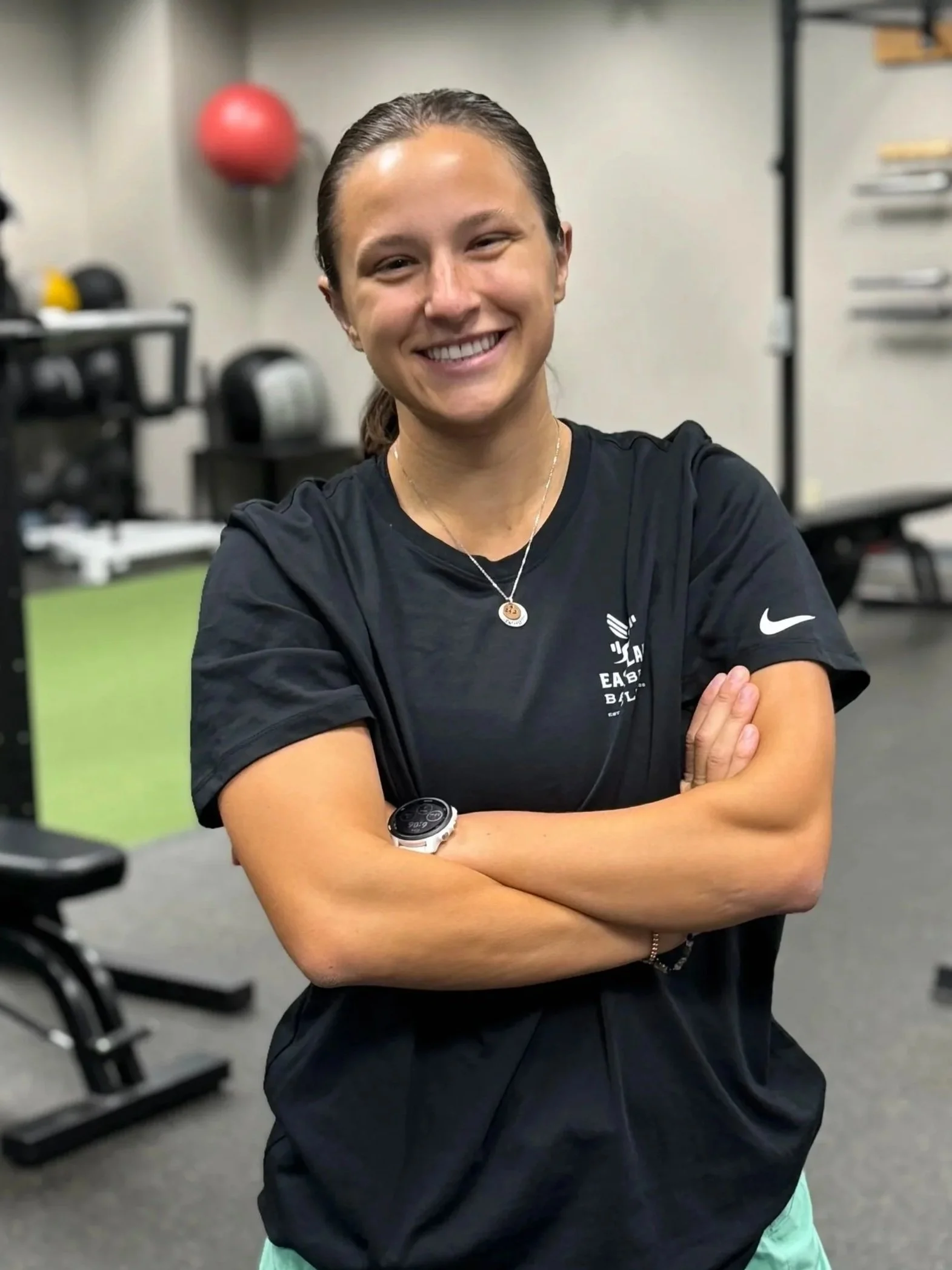 A woman smiling in a gym, wearing a black T-shirt with athletic logos, with arms crossed.