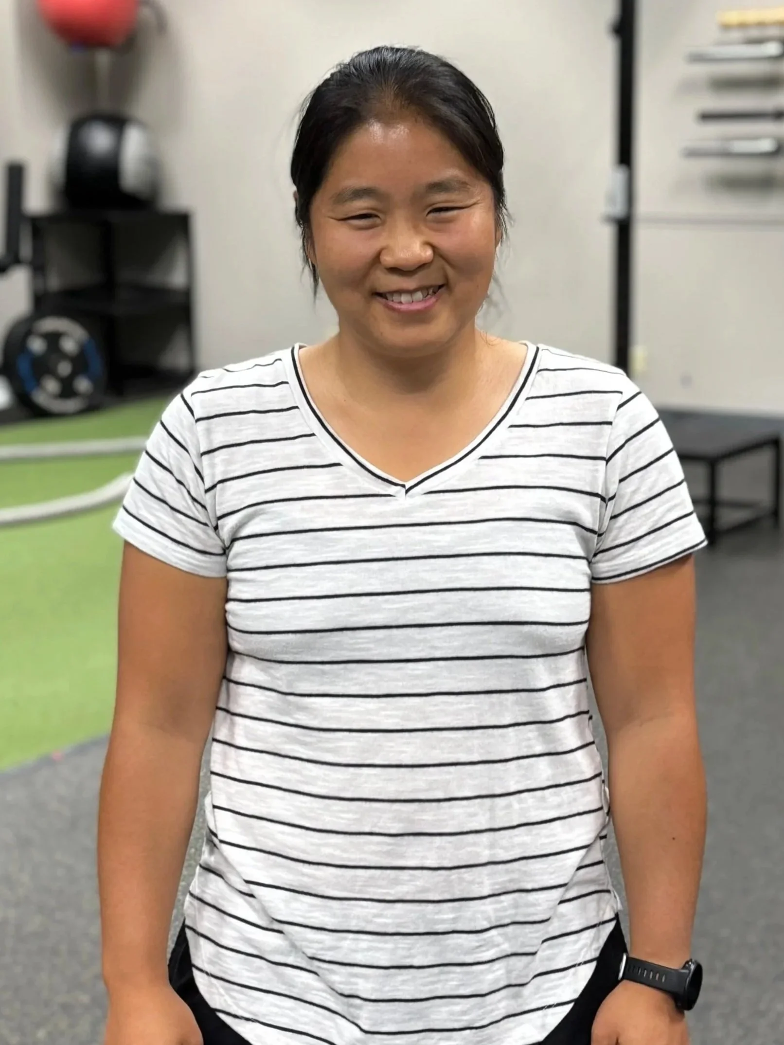A smiling woman with black hair in a striped white and black t-shirt standing in a gym.