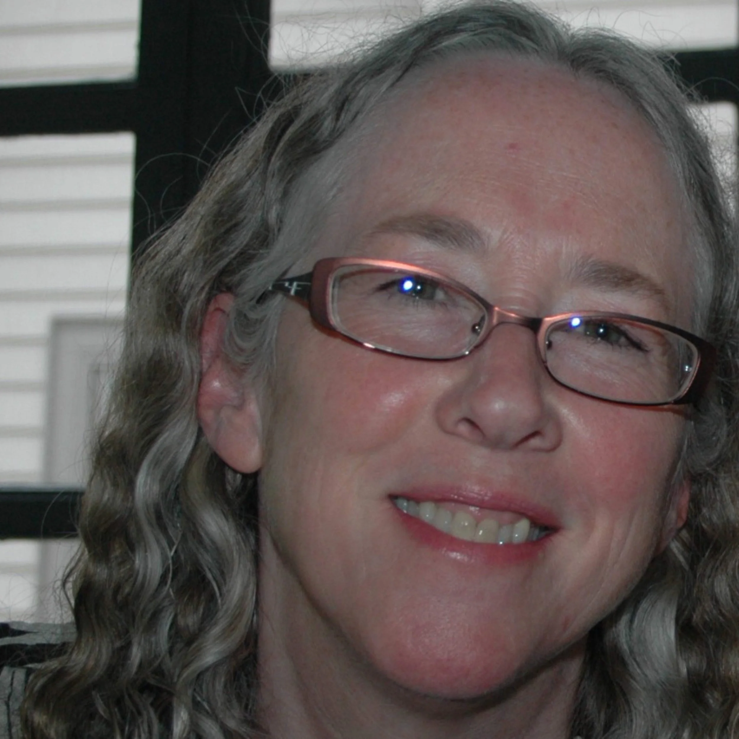 Close-up of a smiling woman with curly gray hair and glasses, indoors near window blinds.