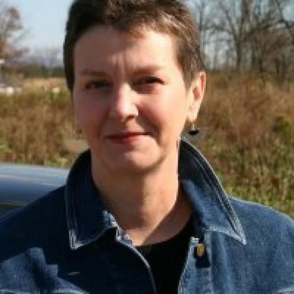 A person with short brown hair wearing a denim jacket outdoors during daytime, with trees and a field in the background.