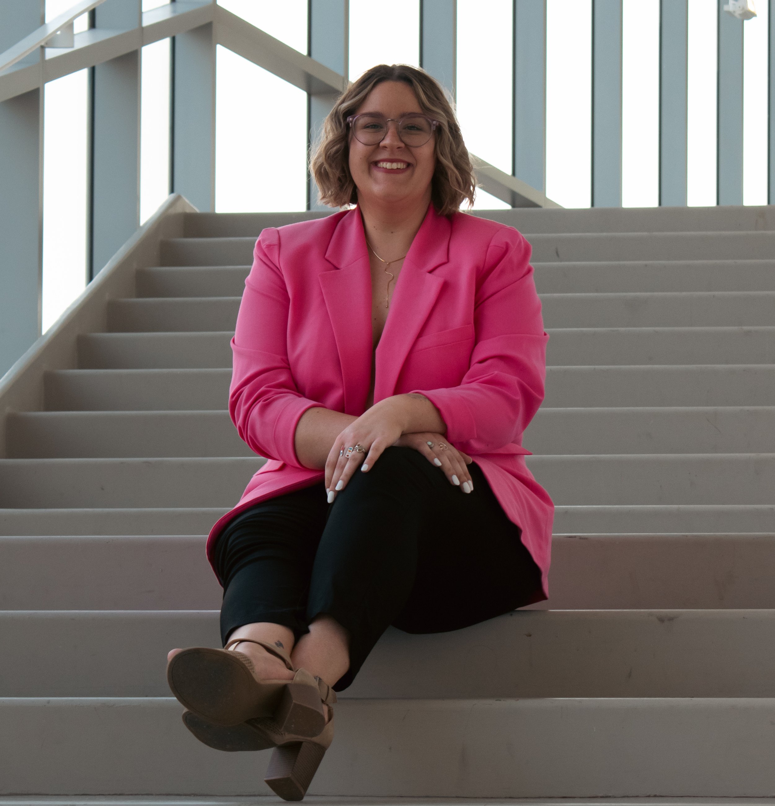 A woman with shoulder-length wavy hair, glasses, and a pink blazer sitting casually on a staircase with large windows in the background, smiling at the camera.