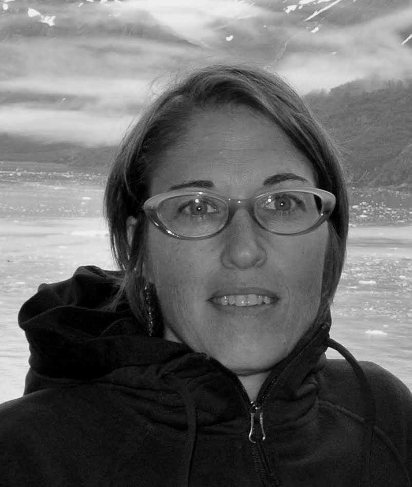 A woman with glasses wearing a black hoodie outdoors with a landscape of open water, mountains, and sky in the background.