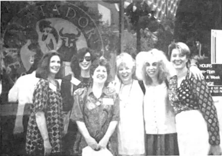 Group of six women standing together outside a building with a sign behind them.
