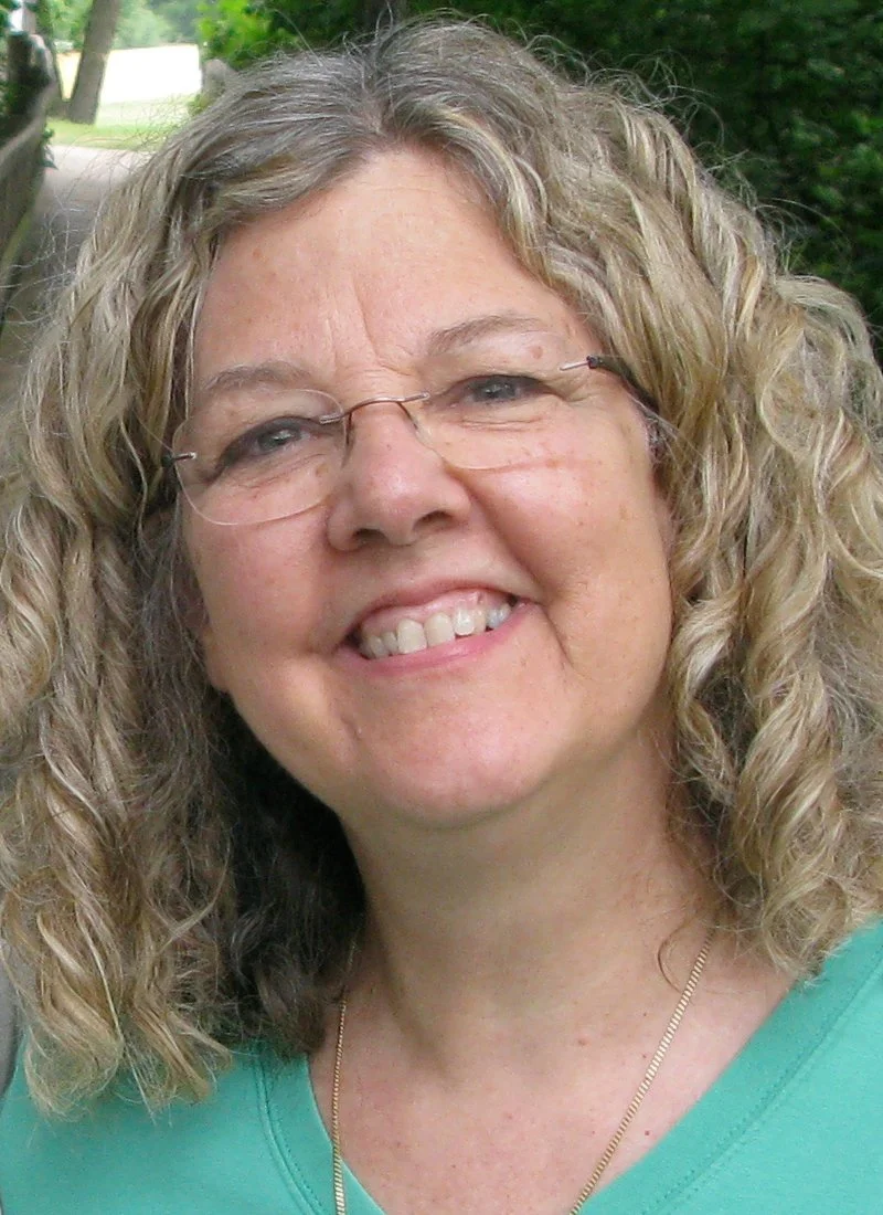 Close-up photo of a woman with curly blonde hair, glasses, and a bright smile, wearing a turquoise top and a thin gold necklace outdoors.