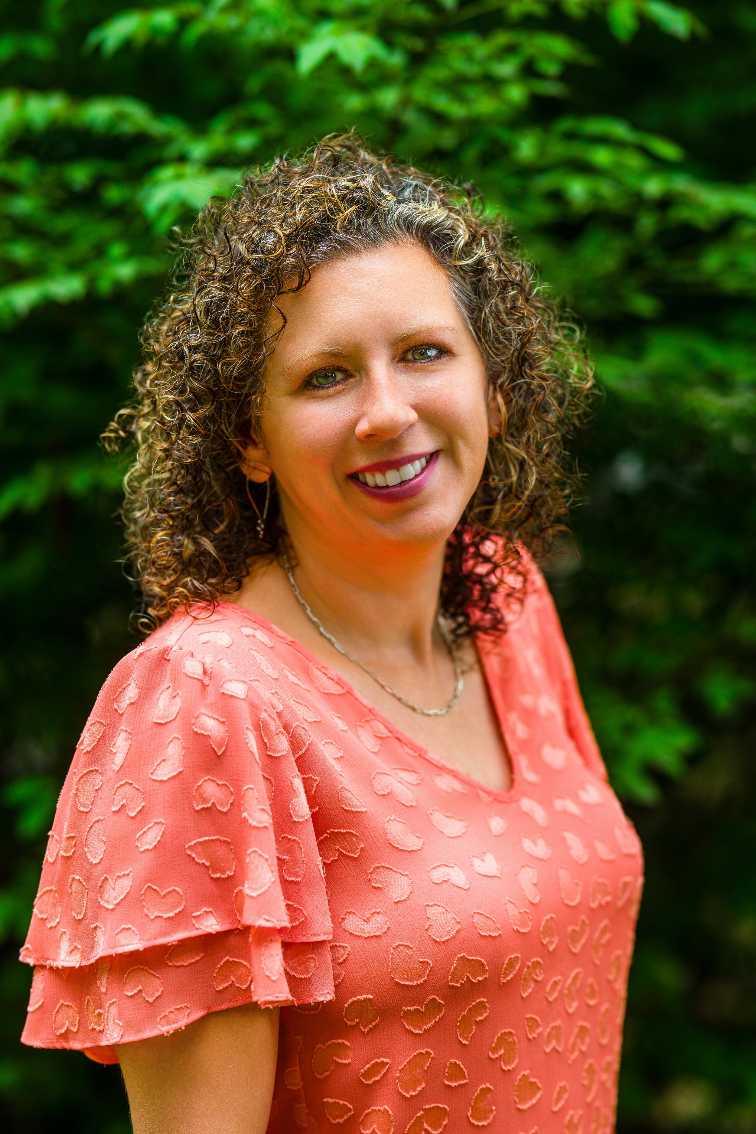 A woman with curly hair wearing a coral-colored top with heart shapes, smiling outdoors with green foliage in the background.