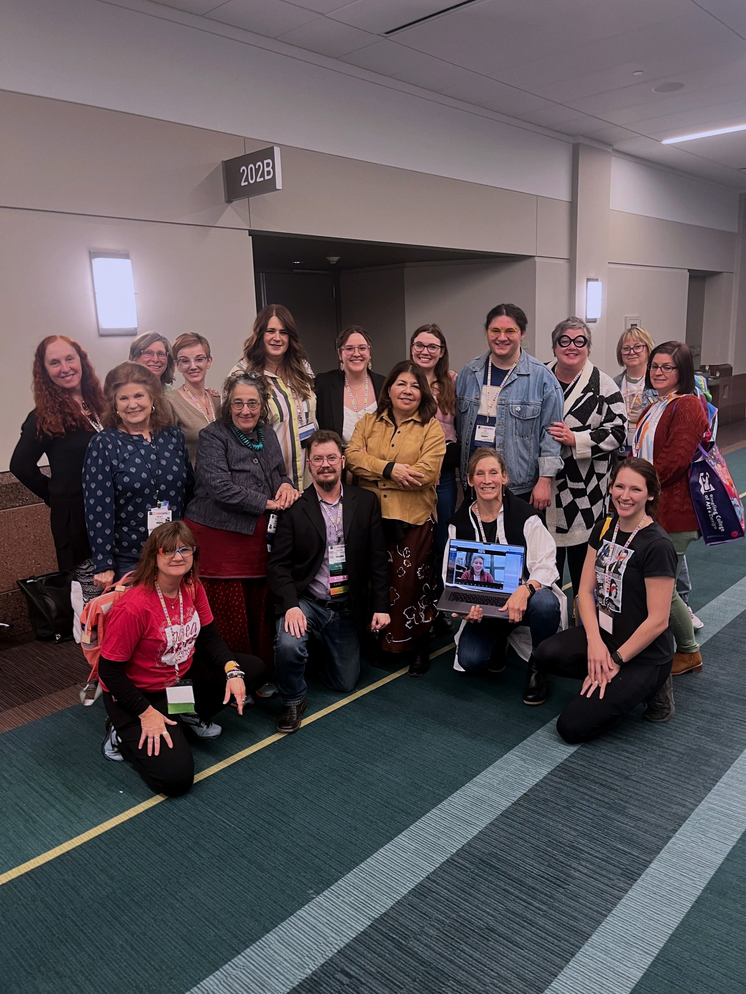 Group of people gathered together in a conference hallway, some kneeling and some standing, with several wearing conference badges. There is a laptop displaying the group in the foreground.