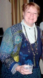 A middle-aged woman smiling, holding a glass, standing indoors with a curtain in the background, wearing a colorful patterned jacket.