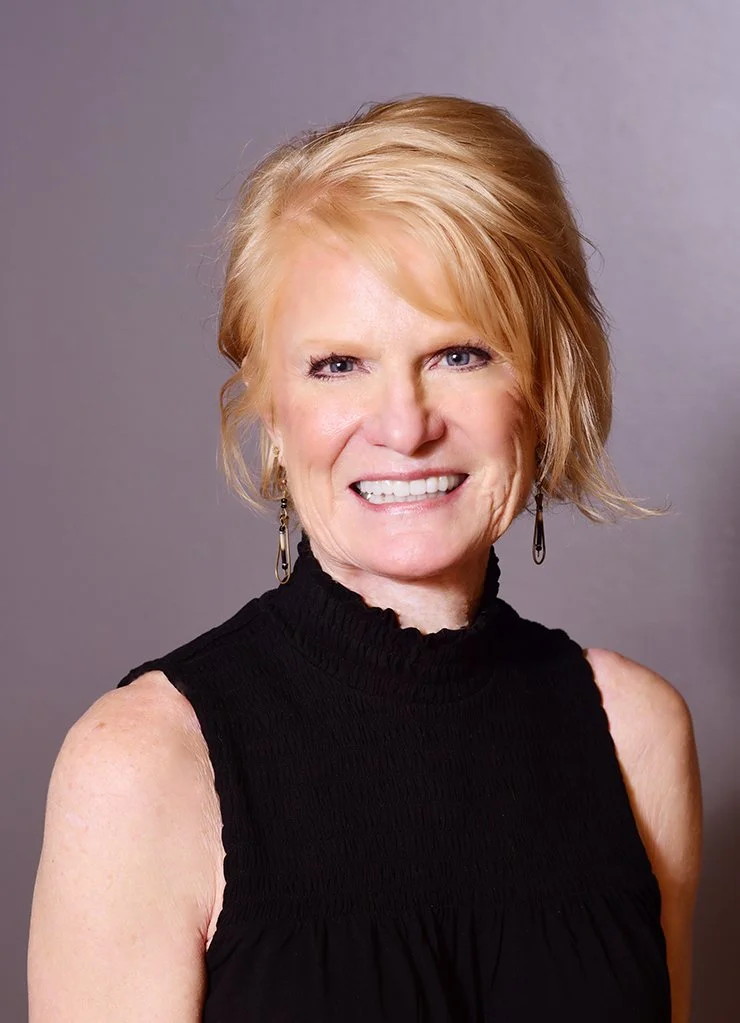 A woman with short, wavy blonde hair smiling, wearing a black sleeveless turtleneck top and dangling earrings, against a gray background.