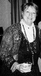 An elderly woman with short hair holding a drink in one hand, wearing a patterned top and necklaces, smiling at the camera.