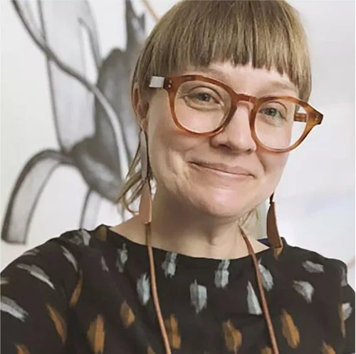 A woman with short light brown hair and glasses smiling at the camera, standing in front of a wall with a mural of a horse.
