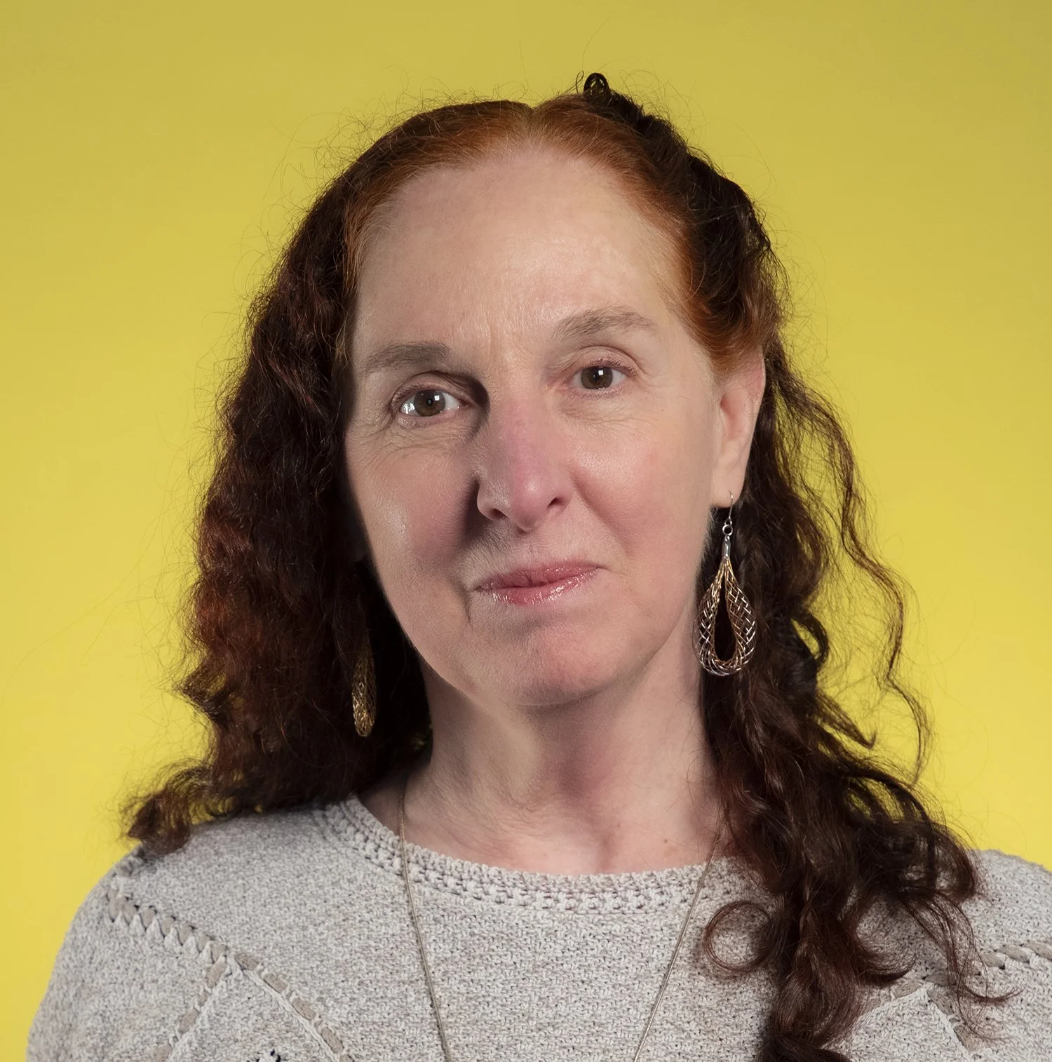 A woman with curly red hair and earrings against a yellow background.