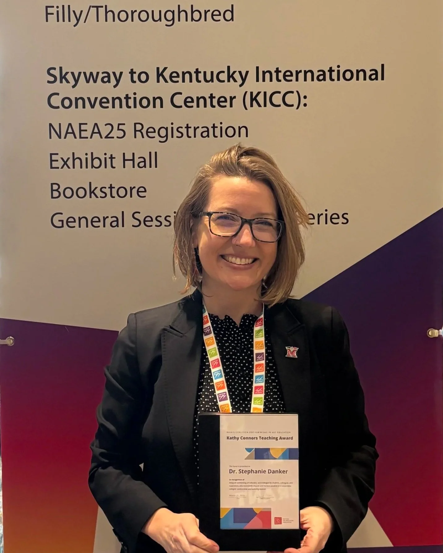 Smiling woman with glasses wearing a black blazer and polka dot shirt, holding a teaching award plaque, standing in front of a conference sign with a gradient background from maroon to purple, displaying event details for Kentucky International Convention Center.