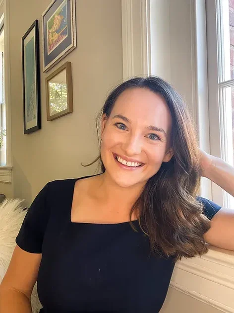 A woman with shoulder-length brown hair smiling and sitting by a window in a room decorated with framed pictures.