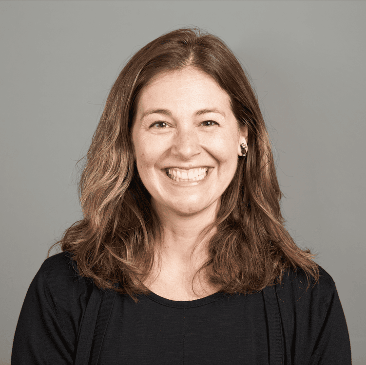 A woman with shoulder-length brown hair smiling, wearing a black top, against a light gray background.