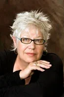 Portrait of an older woman with short, spiky white hair and glasses, wearing a black top, resting her chin on her hand.