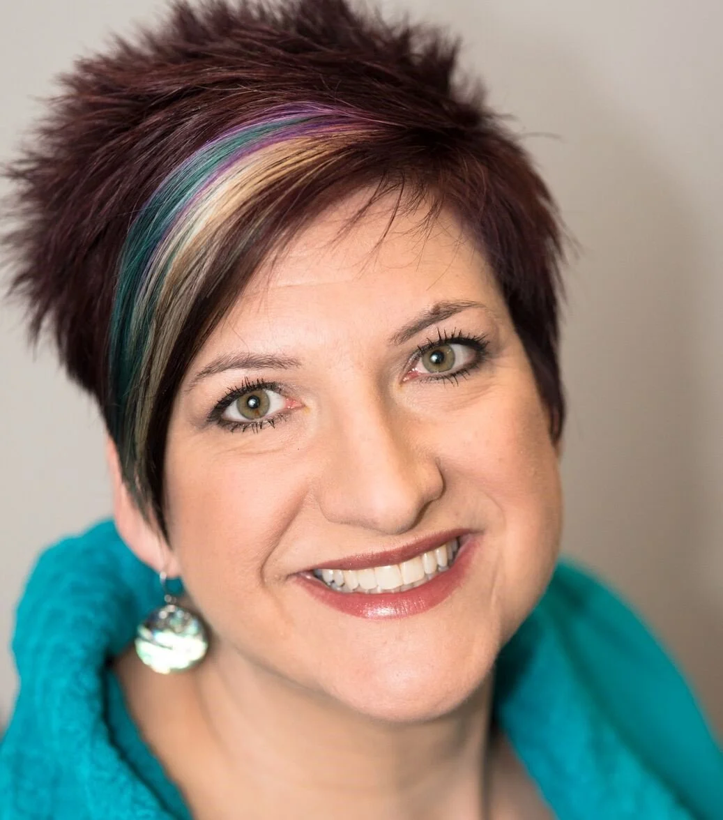 A woman with short spiky hair, dyed with rainbow-colored streaks, smiling and wearing a turquoise sweater and silver earrings.