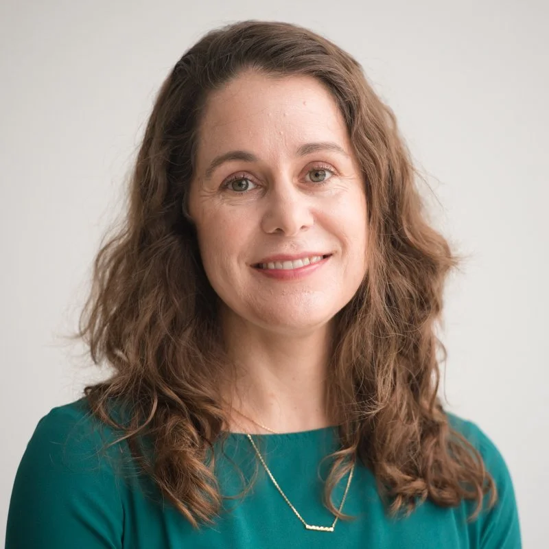 Portrait of a woman with shoulder-length wavy brown hair, wearing a green top and a delicate necklace, smiling at the camera against a light background.