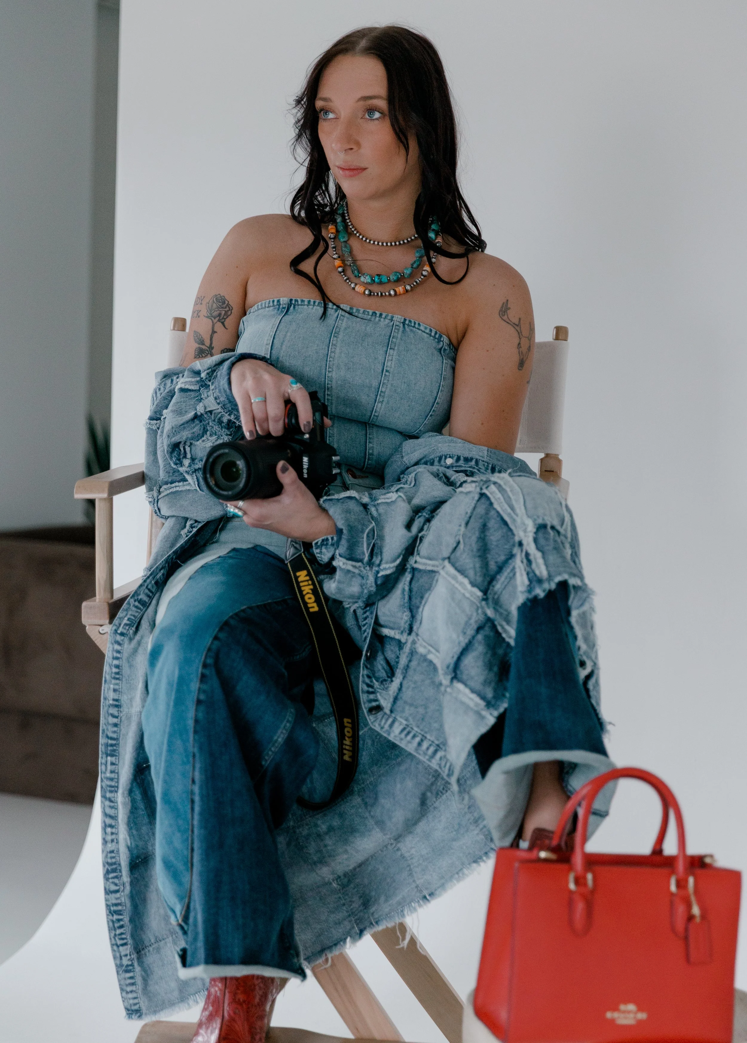 A woman with black hair and tattoos on her arms sitting on a director's chair holding a Nikon camera. She is wearing a strapless denim top, layered necklaces, and jeans with a patchwork design. In front of her is a red handbag.