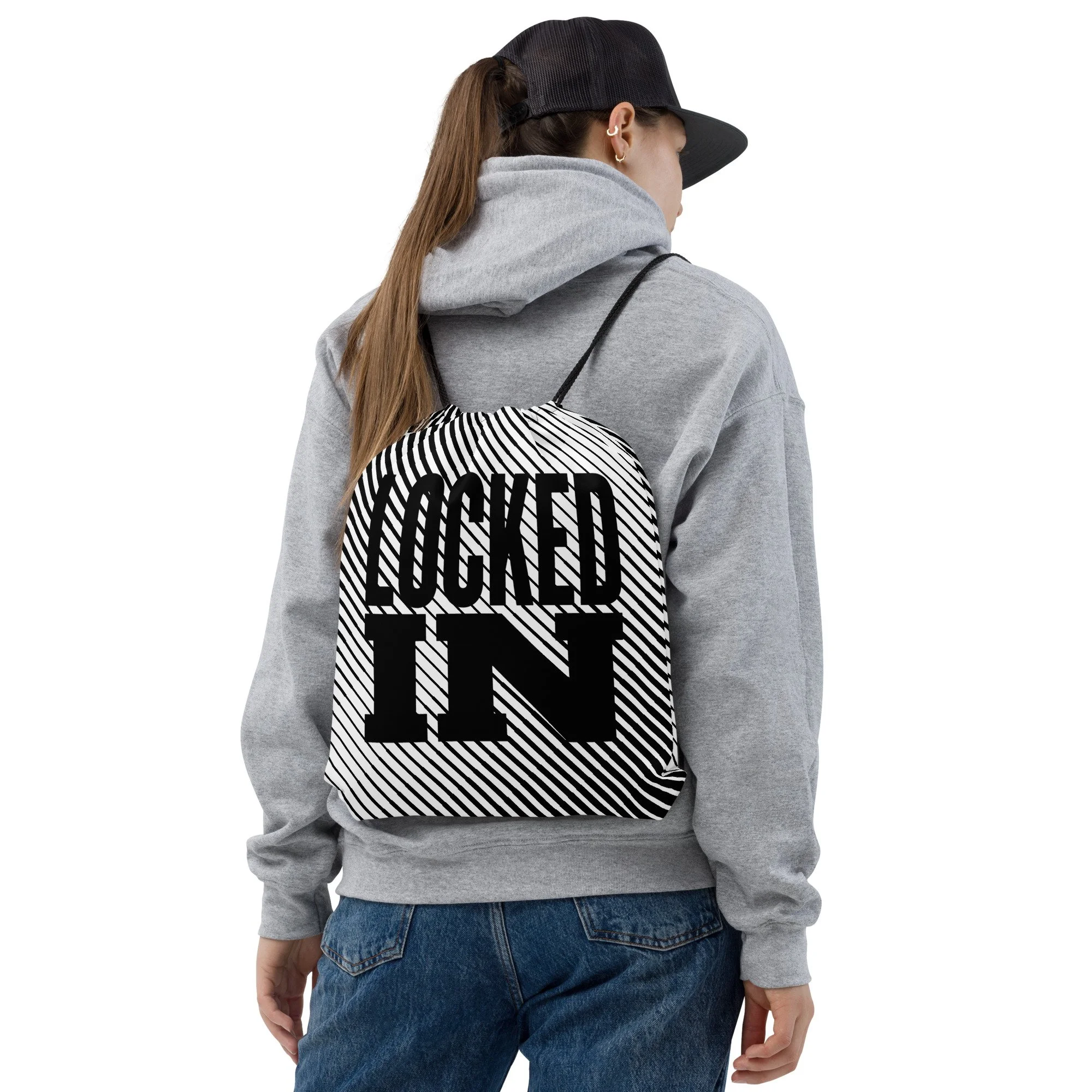 A person with long hair wearing a black baseball cap, gold hoop earrings, a light gray hoodie, and blue jeans, standing against a white background.