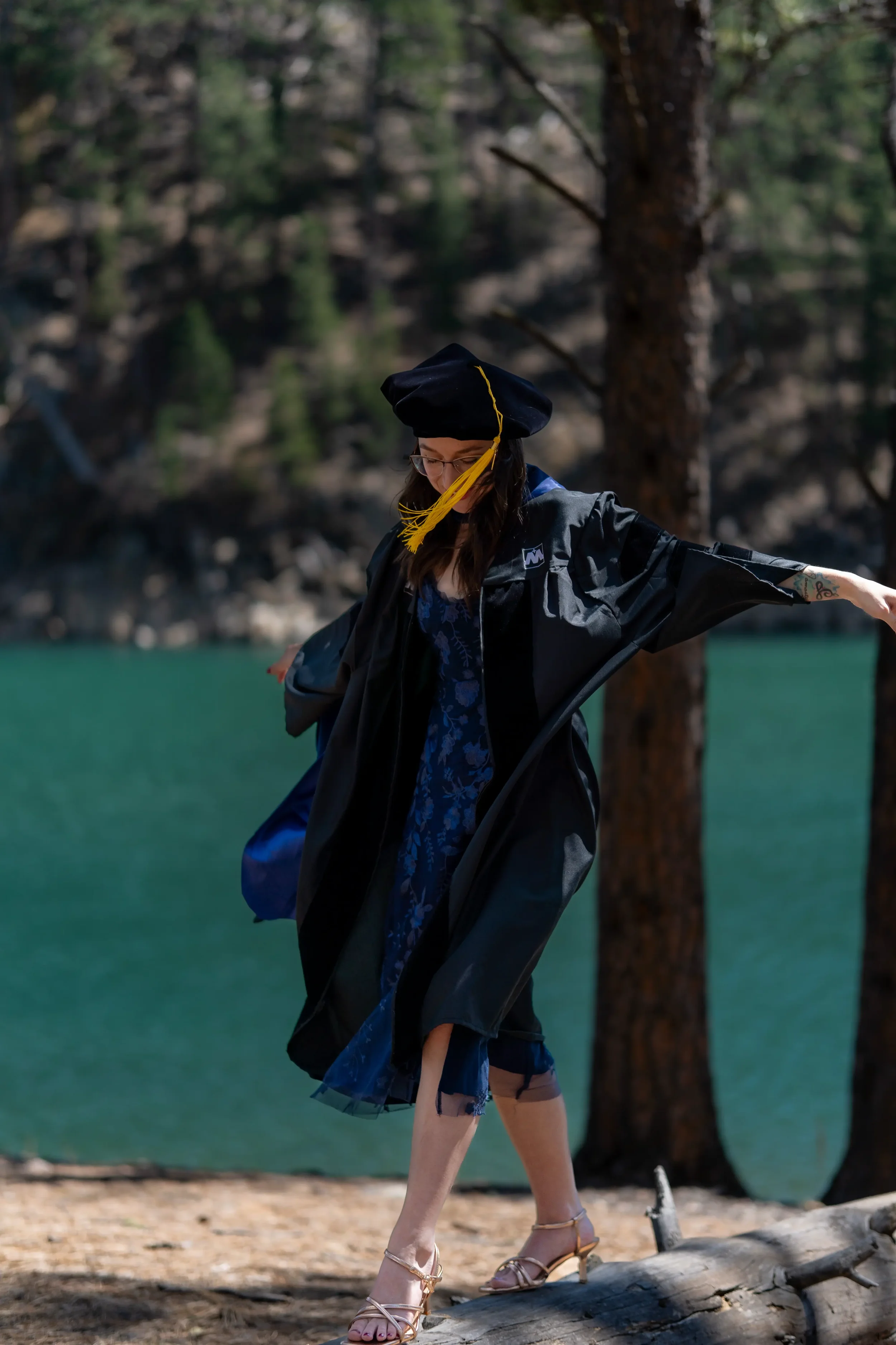 A woman in a graduation cap and gown standing on a rocky surface outdoors with trees and water in the background