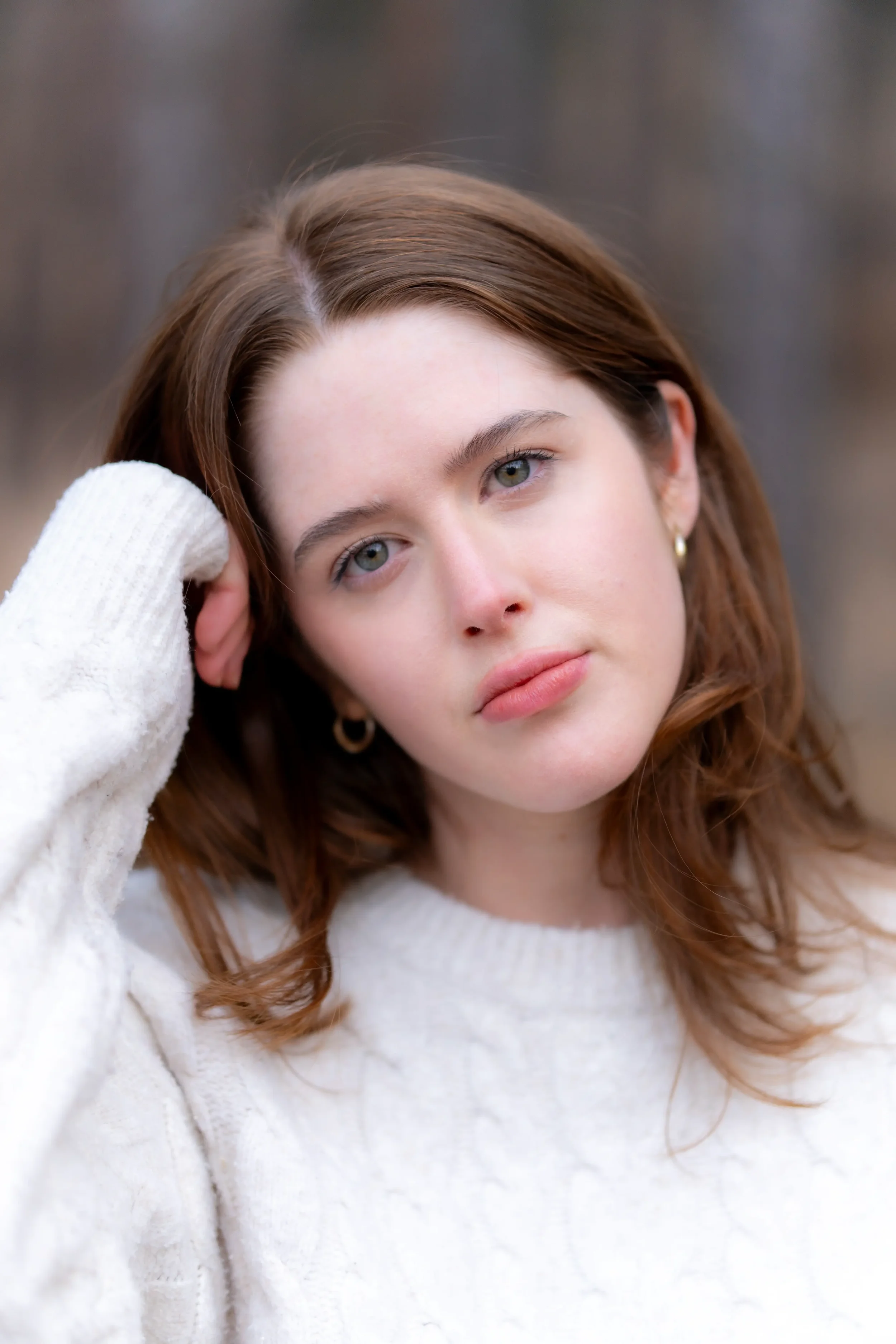 Close-up of a young woman with green eyes and brown hair, wearing a white sweater, resting her head on her hand outdoors.