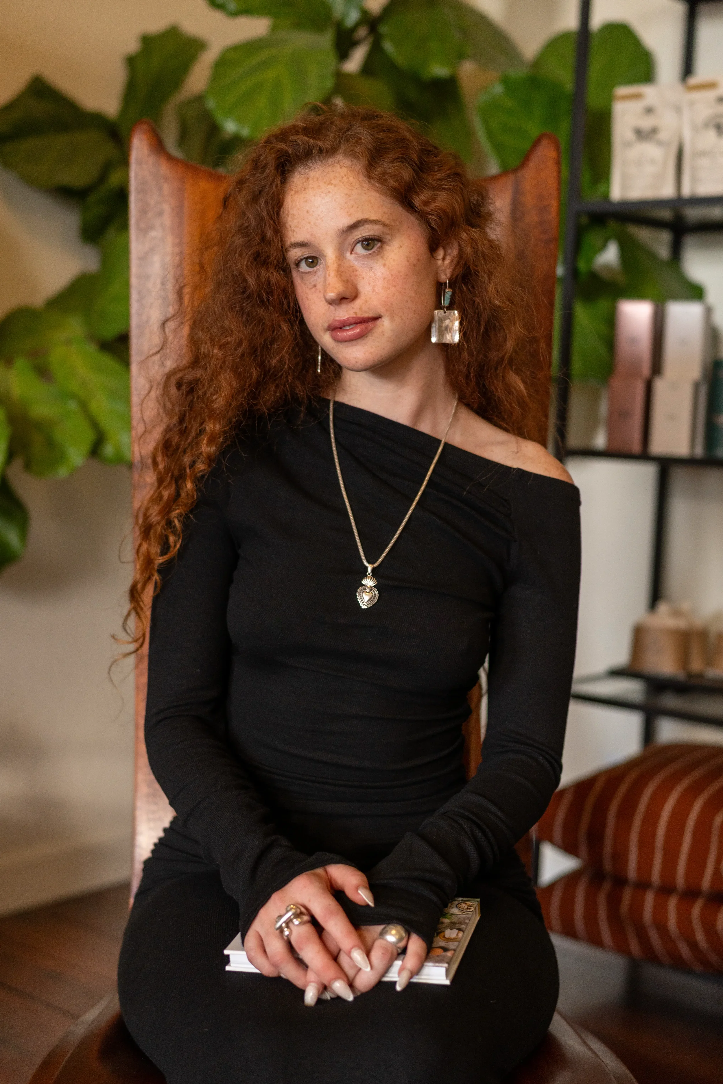 A young woman with curly red hair, wearing a black long-sleeve top and large earrings, sitting on a chair in a cozy room with plants and bookshelves.