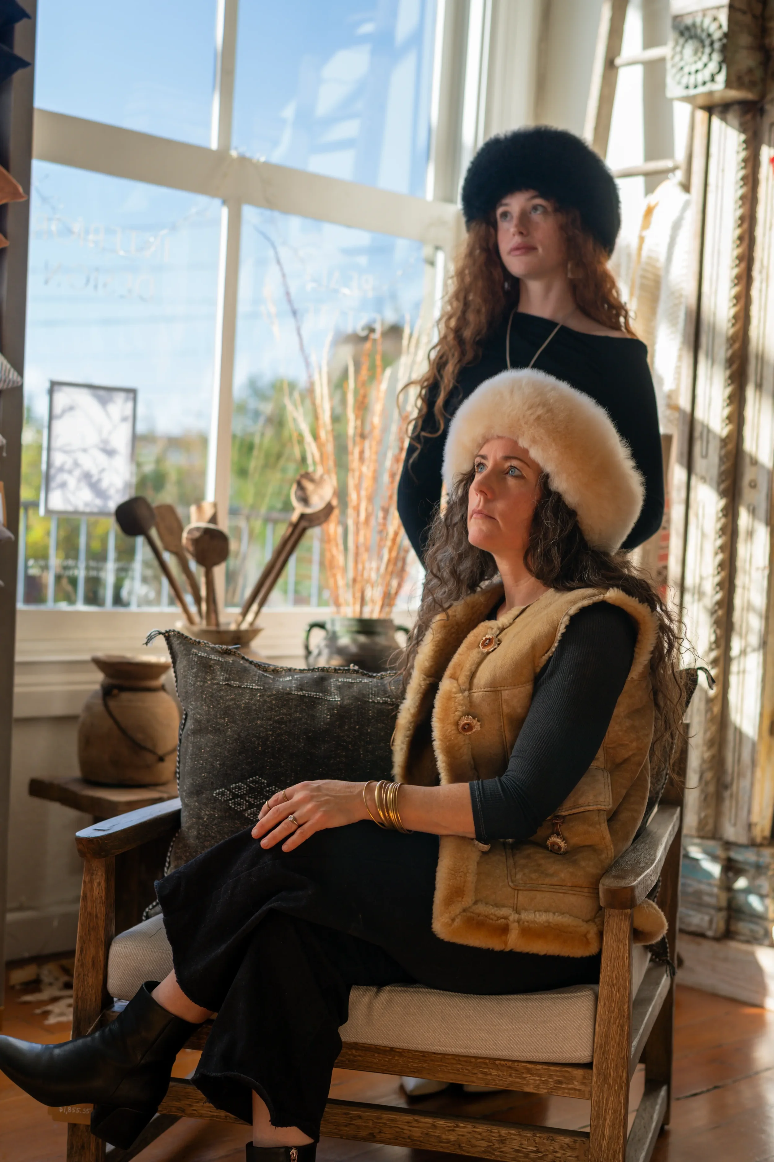 Two women wearing fur hats indoors, one sitting on a wooden bench and the other standing behind her, with large windows and wooden decor in the background.