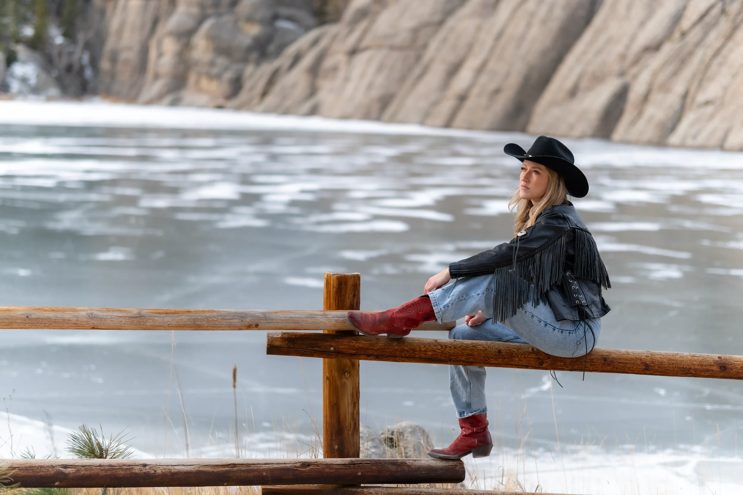 A woman wearing a black cowboy hat, black fringed jacket, blue jeans, and red cowboy boots is sitting on a wooden railing by a body of water with a rocky shoreline in the background.