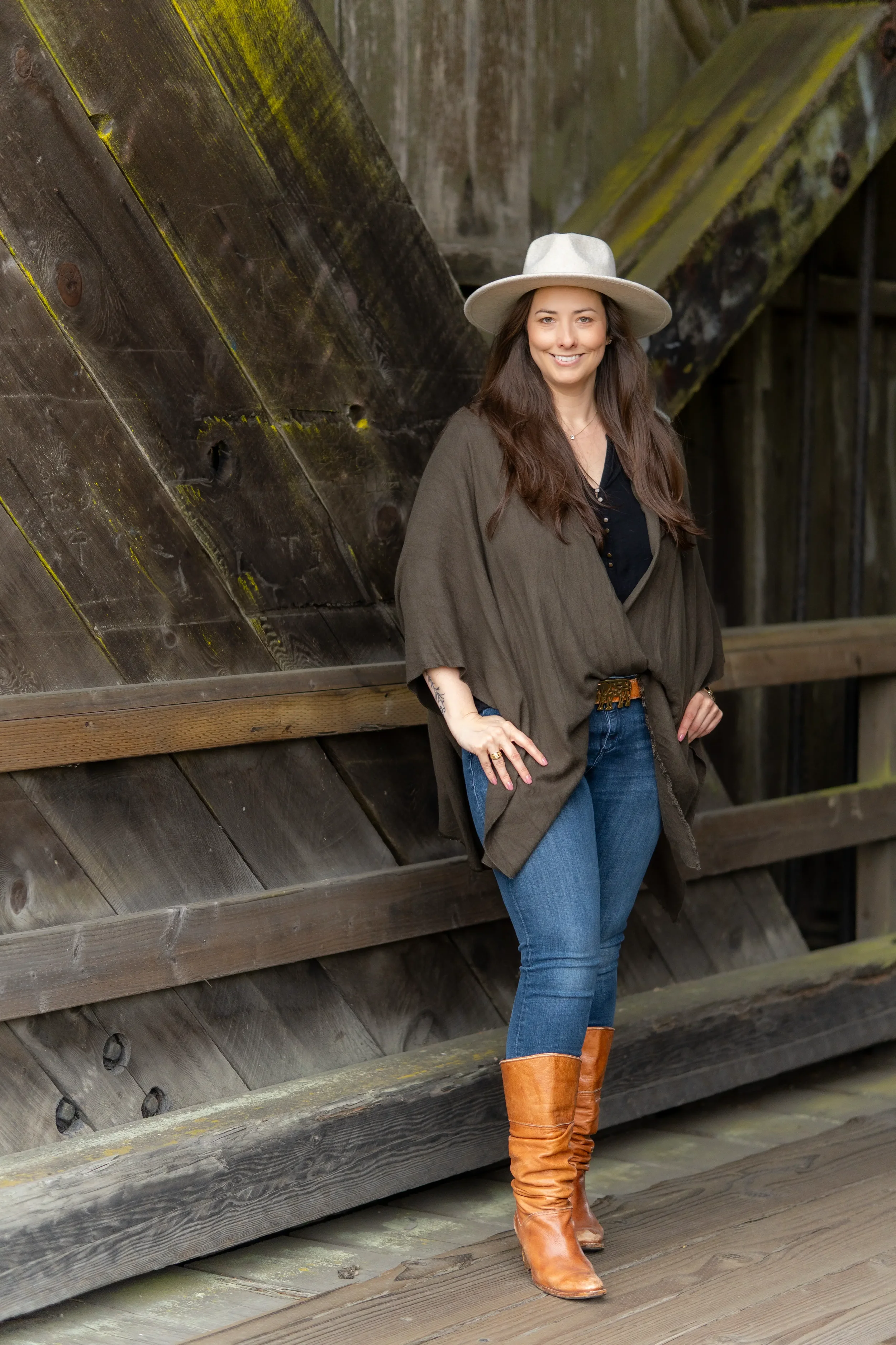 A woman with long brown hair wearing a wide-brimmed beige hat, a dark top, an olive-green jacket, and blue jeans, standing against a weathered wooden structure with a smile.