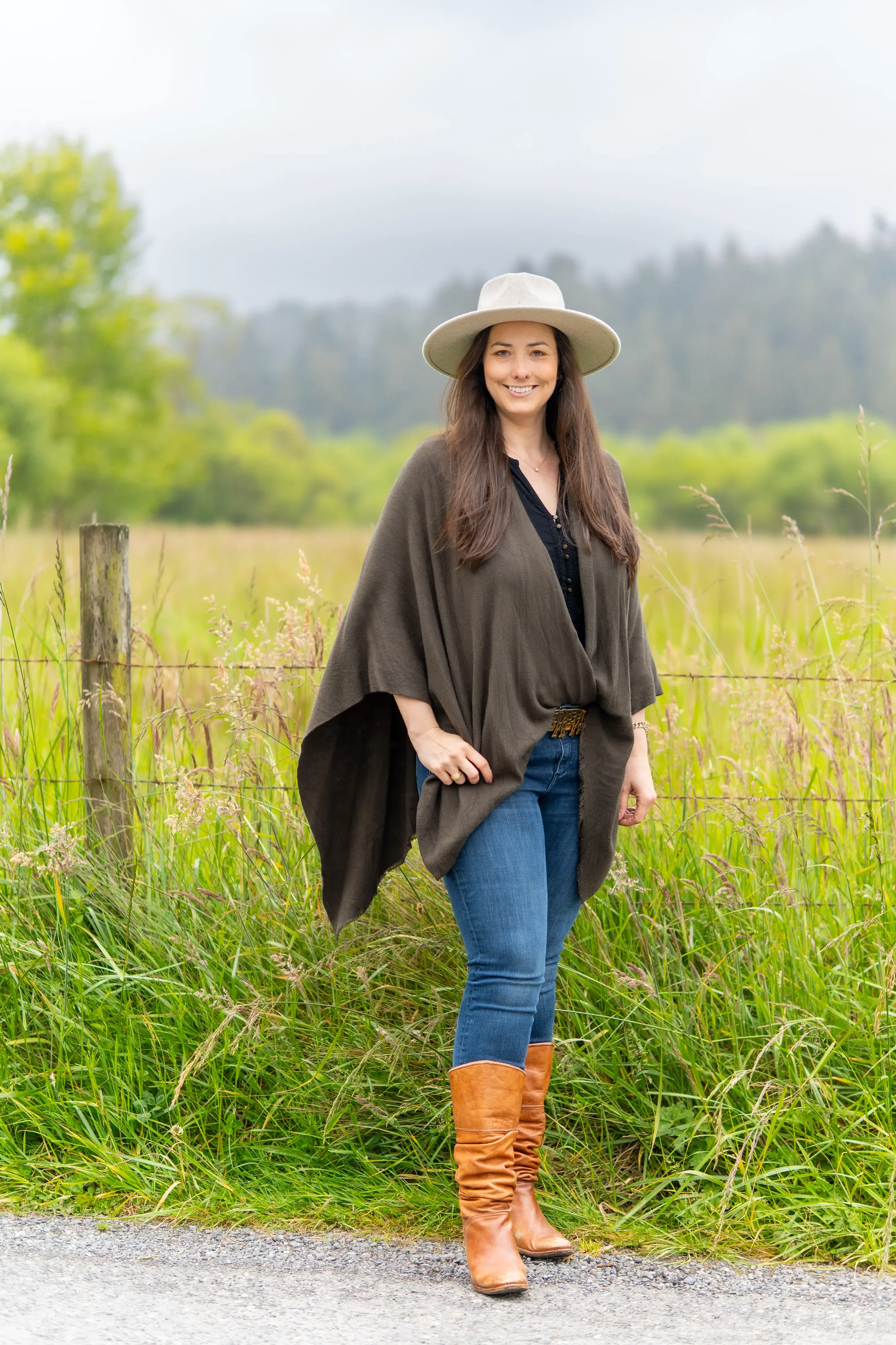A woman with long brown hair, wearing a wide-brimmed hat, dark top, gray shawl, blue jeans, and tan boots, standing outdoors on a gravel path with green grass and trees in the background.